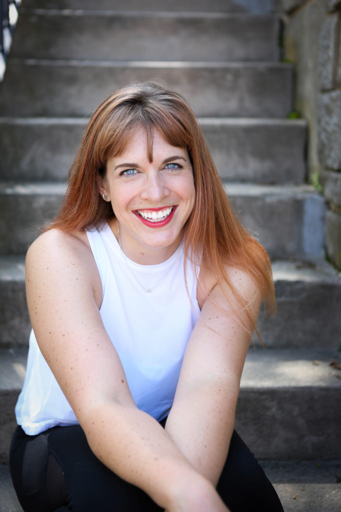 Jenna is wearing a white tank top and black leggings. She's sitting on a concrete staircase and smiling.