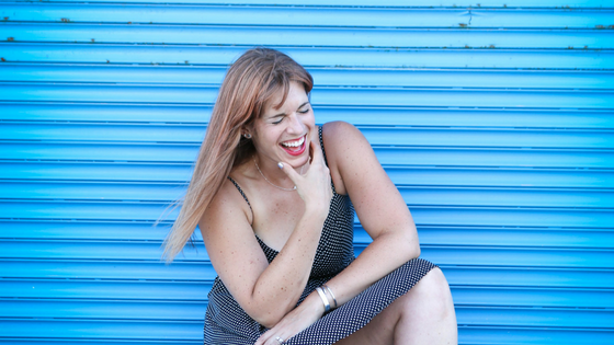 Jenna is crouched down and laughing. There is a blue wall in the background and she's wearing a polka dot spaghetti strap dress and pink converse.