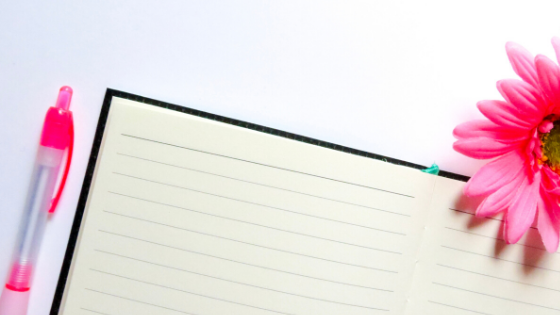 White background with a black and white notebook, a pink pen and a pink gerber daisy.