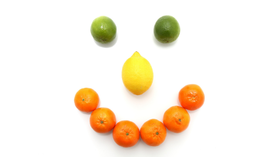 A citrus fruit smiley face on a white background with oranges for a mouth, a lemon nose, and limes for eyes.