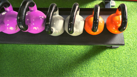 Green turf with 6 fuschia, gray, and orange kettlebells sitting on a black shelf.