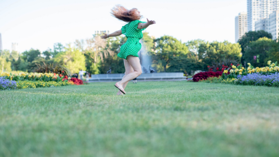 A girl with light brown hair is wearing a green romper and twirling in the park. The Chicago skyline is in the background.