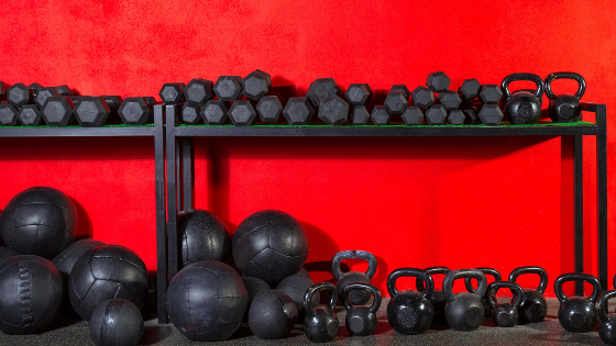 A rack of black dumbbells, medicine balls, and kettlebells against a red wall.