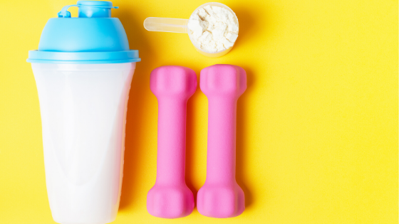 White shaker bottle with a blue lid, little pink dumbbells, and a scoop of white protein powder on a bright yellow background.