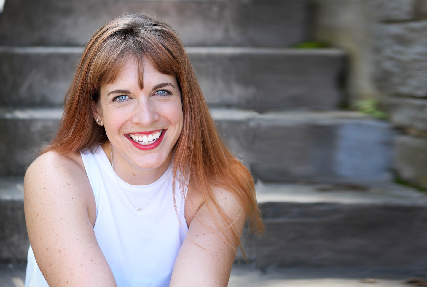 Jenna is wearing a white tank top and black leggings. She's sitting on a concrete staircase and smiling.