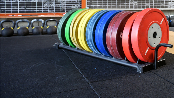 Rainbow colored barbell plates on a rack in a gym. The floor is black and there are chalkboards and kettlebells in the background.