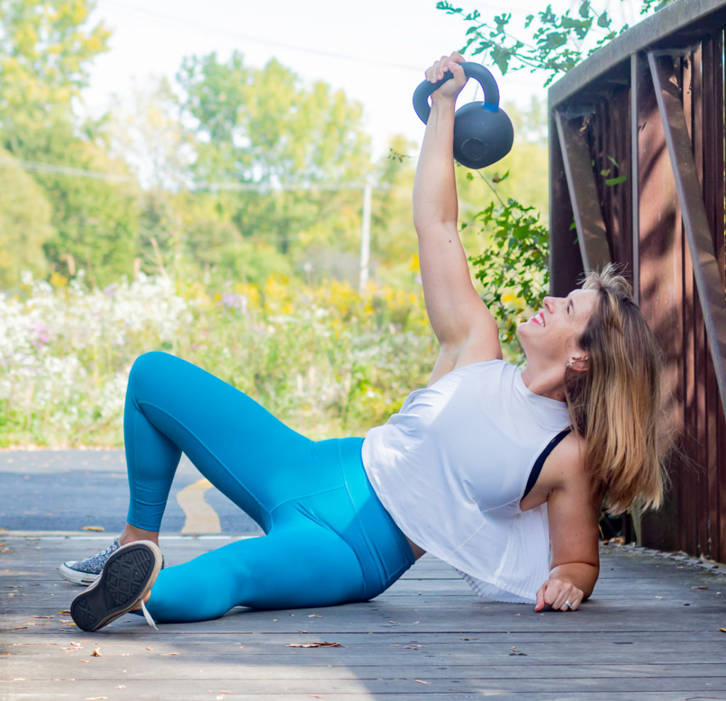 Jenna is wearing silver glitter converse, teal leggings and a white top. She is sitting on a bridge doing a half-Turkish getup, holding a black kettlebell overhead. There is a green field with wildflowers in the background.