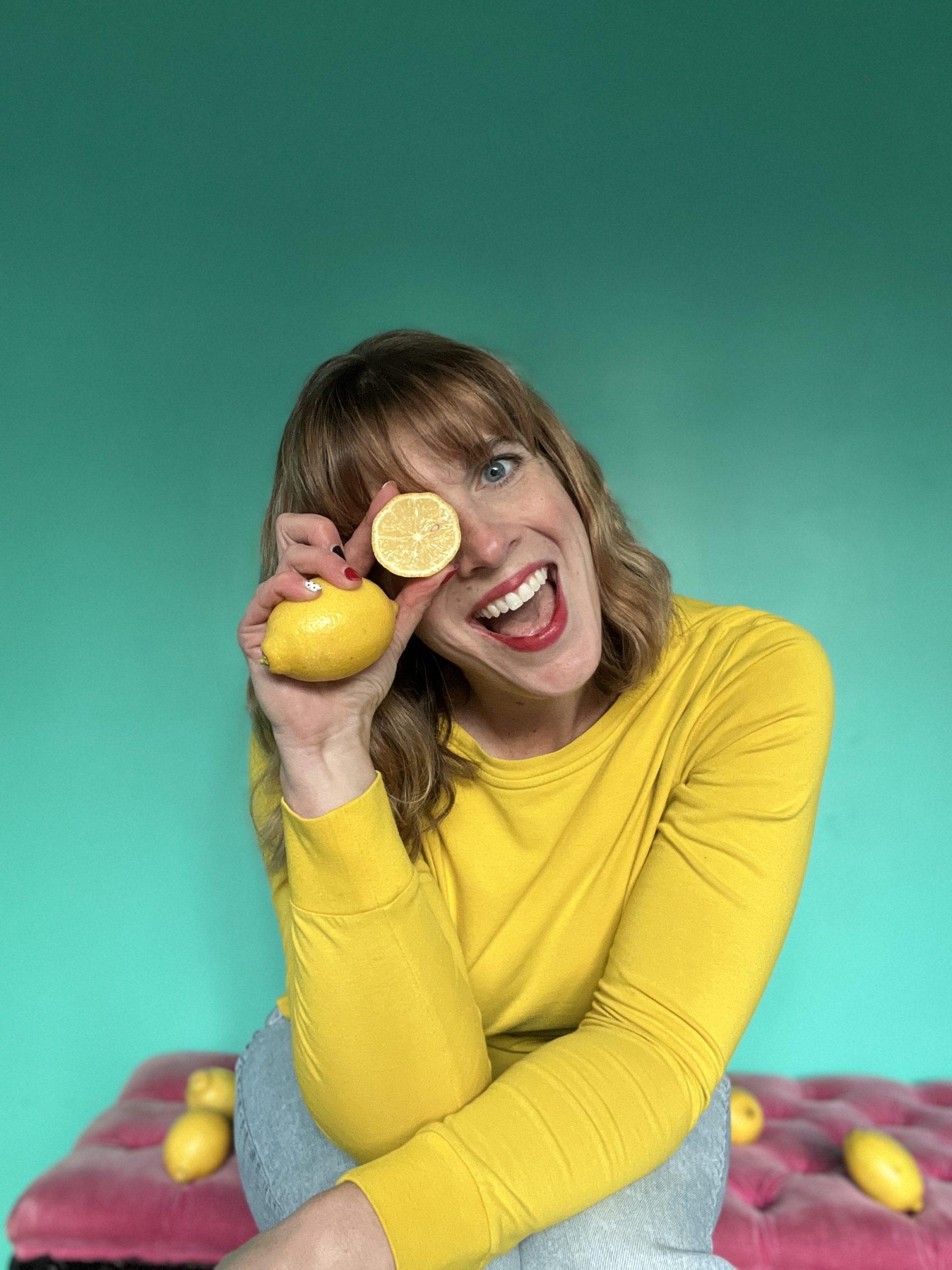 Jenna is wearing a bright yellow long sleeved shirt with light blue jeans. She is sitting on a pink bench with a turquoise backdrop. There are 4 lemons on the bench and she is holding them with one over her eye. Her head is tilted to the side and she is smiling with an open mouth.