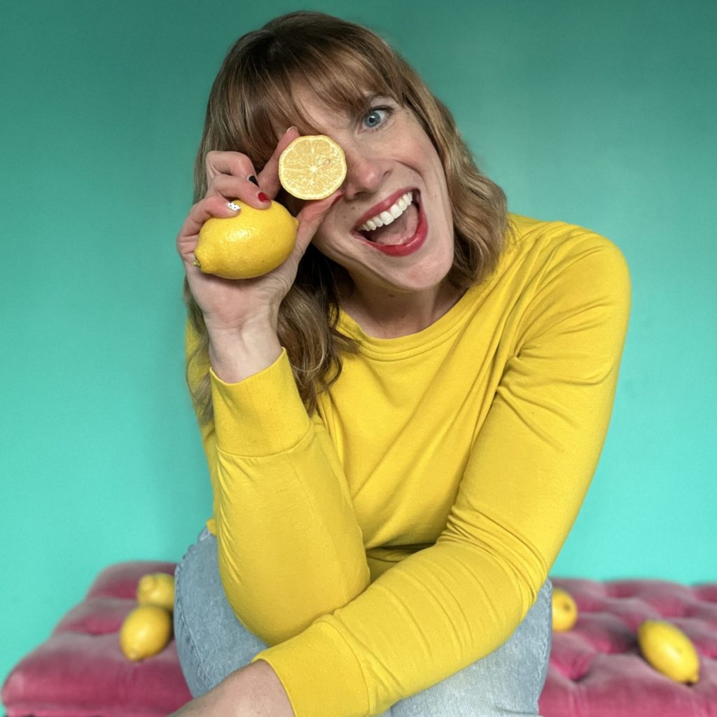 Jenna is wearing a bright yellow long sleeved shirt with light blue jeans. She is sitting on a pink bench with a turquoise backdrop. There are 4 lemons on the bench and she is holding them with one over her eye. Her head is tilted to the side and she is smiling with an open mouth.