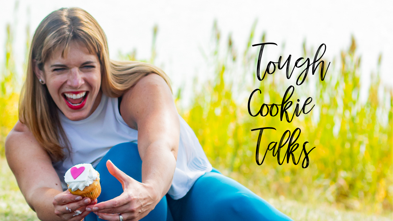 Jenna is wearing bright blue leggings and a white shirt. She is squatting down on the sidewalk holding a cupcake and laughing. There is tall green grass and a lake in the background. The words Tough Cookie Talks are in black type to her right.